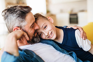 La qualité chez Tchibo pour la famille : un père en chemise ouverte en denim est assis sur le canapé avec son fils blotti dans ses bras.