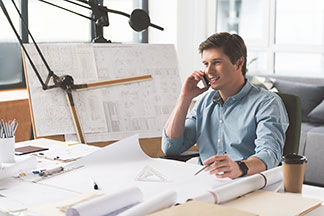Un jeune homme portant une chemise hommes bleue est assis à son bureau, avec une planche à dessin en arrière-plan, et parle sur son smartphone.