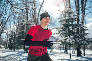 Un jeune homme portant un t-shirt de sport rouge avec, en dessous, un maillot de corps noir manches longues et une casquette noire fait son jogging dehors en hiver.