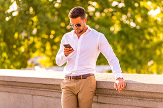 Des chemises manches longues pour le bureau : un homme portant une chemise blanche manches longues et des lunettes de soleil se penche contre la balustrade d'un pont de pierre, en actionnant son smartphone.