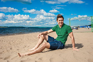 Un jeune homme en short et polo vert s'assoit joyeusement sur la plage sous un soleil radieux.