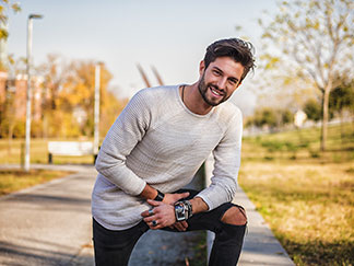 Un jeune homme barbu et souriant se tient dans un parc, vêtu d'un shirt à manches longues de couleur claire.