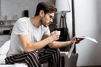 Un jeune homme tenant une tasse de café est assis sur le bord de son lit, en pyjama, et lit le journal.