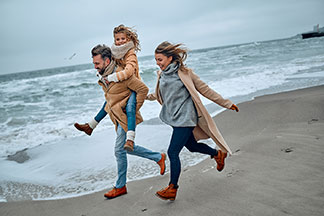 Un homme portant une écharpe et avec sa petite fille sur son dos accompagnés d’une femme marchent se promènent tranquillement le long de la plage. Tous portent des vêtements d'automne.