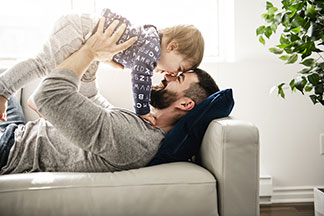Un père en un t-shirt gris manches longues joue sur le canapé avec son fils.