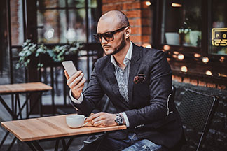 De la qualité pour votre montre hommes : un jeune homme en costume est assis à l'extérieur d'un café et regarde son téléphone portable. Dans son bras, il porte une montre hommes.