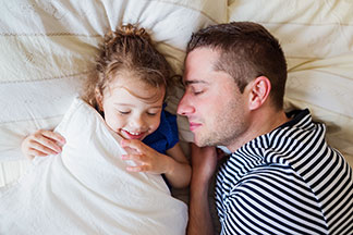 Pas de soucis grâce à la qualité Tchibo : un père en pull rayé s'allonge avec sa fille dans le lit parental et dort.