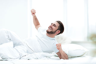 Un jeune homme portant un t-shirt blanc est confortablement allongé le matin, avec encore la moitié de son corps sous sa couverture blanche le matin.