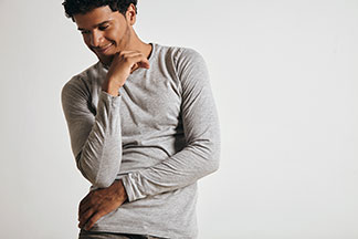 Un jeune homme portant un t-shirt gris manches longues se tient nonchalamment devant un mur gris clair.