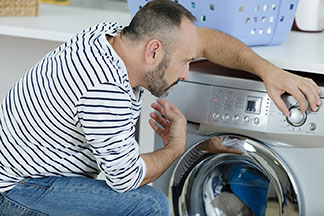 Un homme portant un jean et un pull rayé blanc et noir s'agenouille devant la machine à laver pour laver son pantalon de jogging et règle le programme.