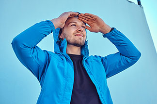 Veste entre saisons pour l'extérieur : Un jeune homme portant une veste Softshell bleue tient ses mains sur son front et regarde au loin.