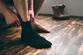 Gros plan sur les jambes d'un homme assis sur son lit et qui met ses bas. Un pied porte déjà une chaussette noire, le deuxième est encore nu.