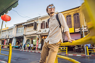 Pantalons hommes pour les loisirs : un jeune homme portant des lunettes de soleil à verres réfléchissants, une chemise en lin crème et des chinos ocres s'appuie avec désinvolture contre une balustrade dans une rue.