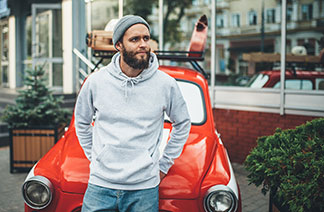 Un jeune homme portant une casquette en laine et un sweat-shirt gris à capuche se penche négligemment sur une voiture rouge.