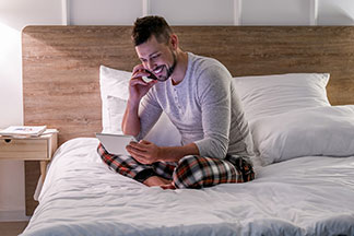 Le soir, un jeune homme est assis sur son lit en pyjama - avec des jambes menottées et un bouton en haut - et parle au téléphone tout en regardant dans sa tablette.