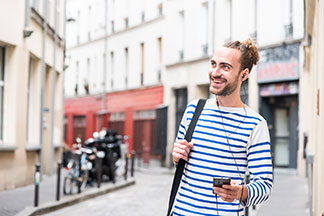 Un jeune homme joyeux, vêtu d'une chemise manches longues rayée bleu et blanc, traverse une ville avec un smartphone à la main.