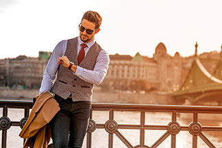 Un homme élégamment vêtu et portant des lunettes de soleil regarde sa montre hommes. Il se tient devant une rivière et s'appuie contre une balustrade.