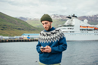 Un jeune homme barbu portant un pull norvégien se tient devant un port, où un navire à passagers vient d'entrer, et regarde son smartphone.