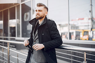 Veste hommes pour le bureau : un jeune homme portant un manteau court noir et une veste grise en dessous est appuyé contre une balustrade, tenant ses lunettes de soleil à la main.
